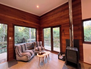 A cozy cabin interior with a sofa, two chairs, a small table, a fireplace, and large windows offering a view of the forest.