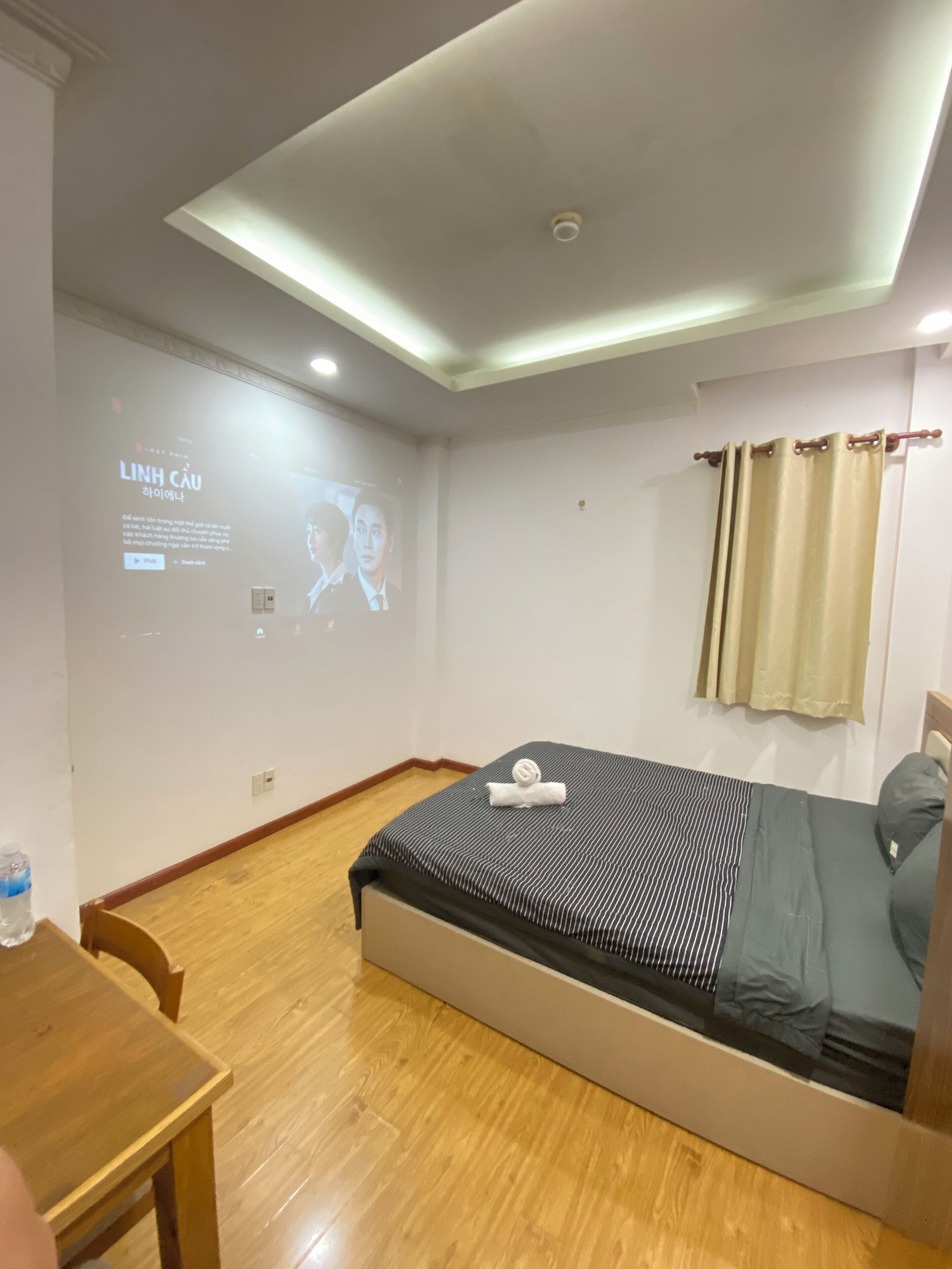 Habitación de hotel con cama, escritorio de madera, pantalla de proyector mostrando una película y cortina.