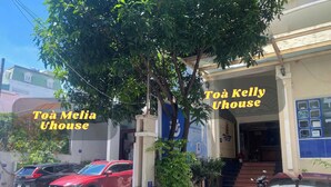 A building with a sign reading "Toà Kelly Uhouse" and "Toà Melia Uhouse". There are cars parked in front and a tree in the foreground. - Uhouse - KELLY Thao Dien Apartment (Ho Chi Minh City)