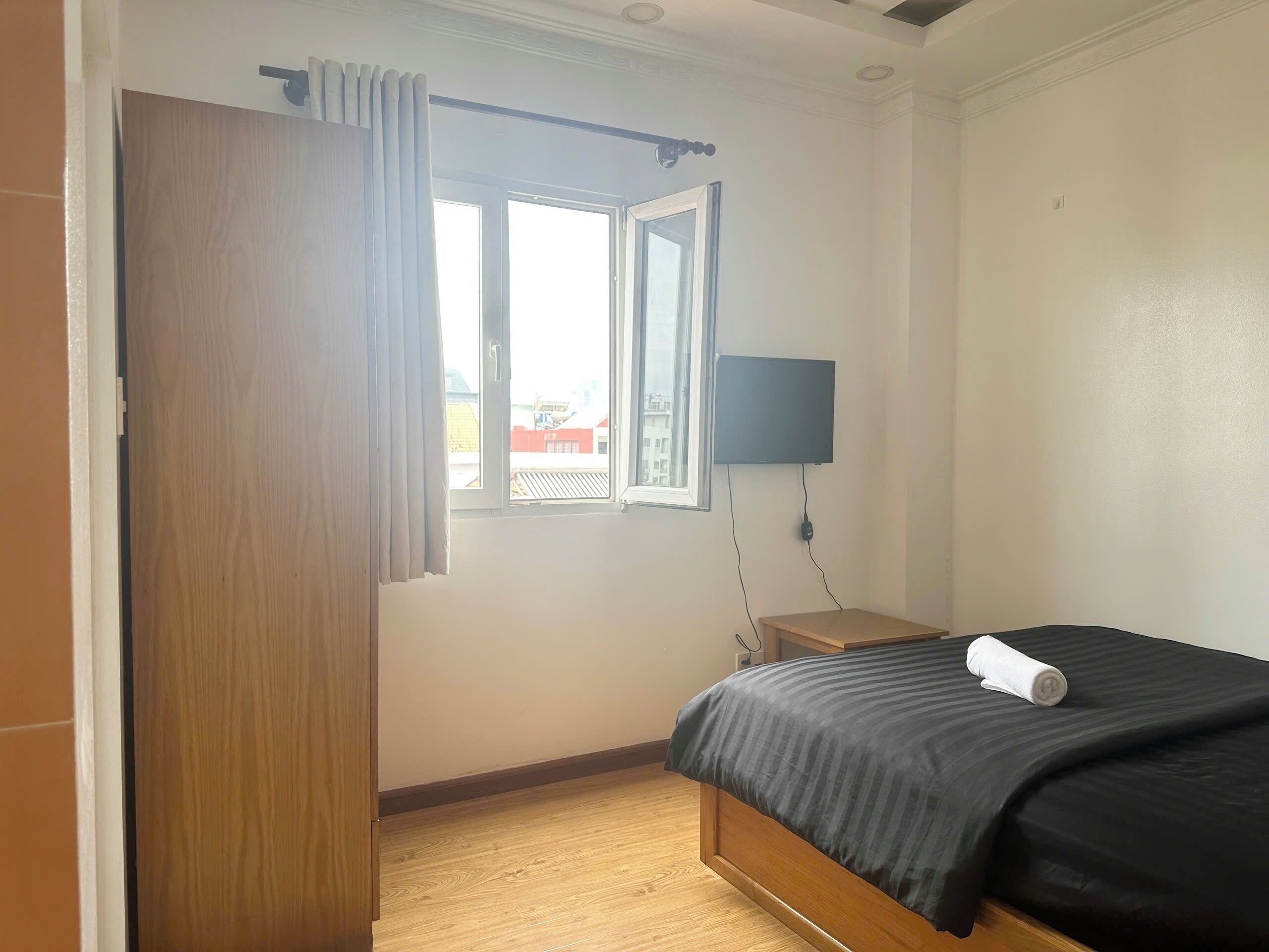 A bedroom with a bed, a window with curtains, a wall-mounted TV, and a wooden nightstand.