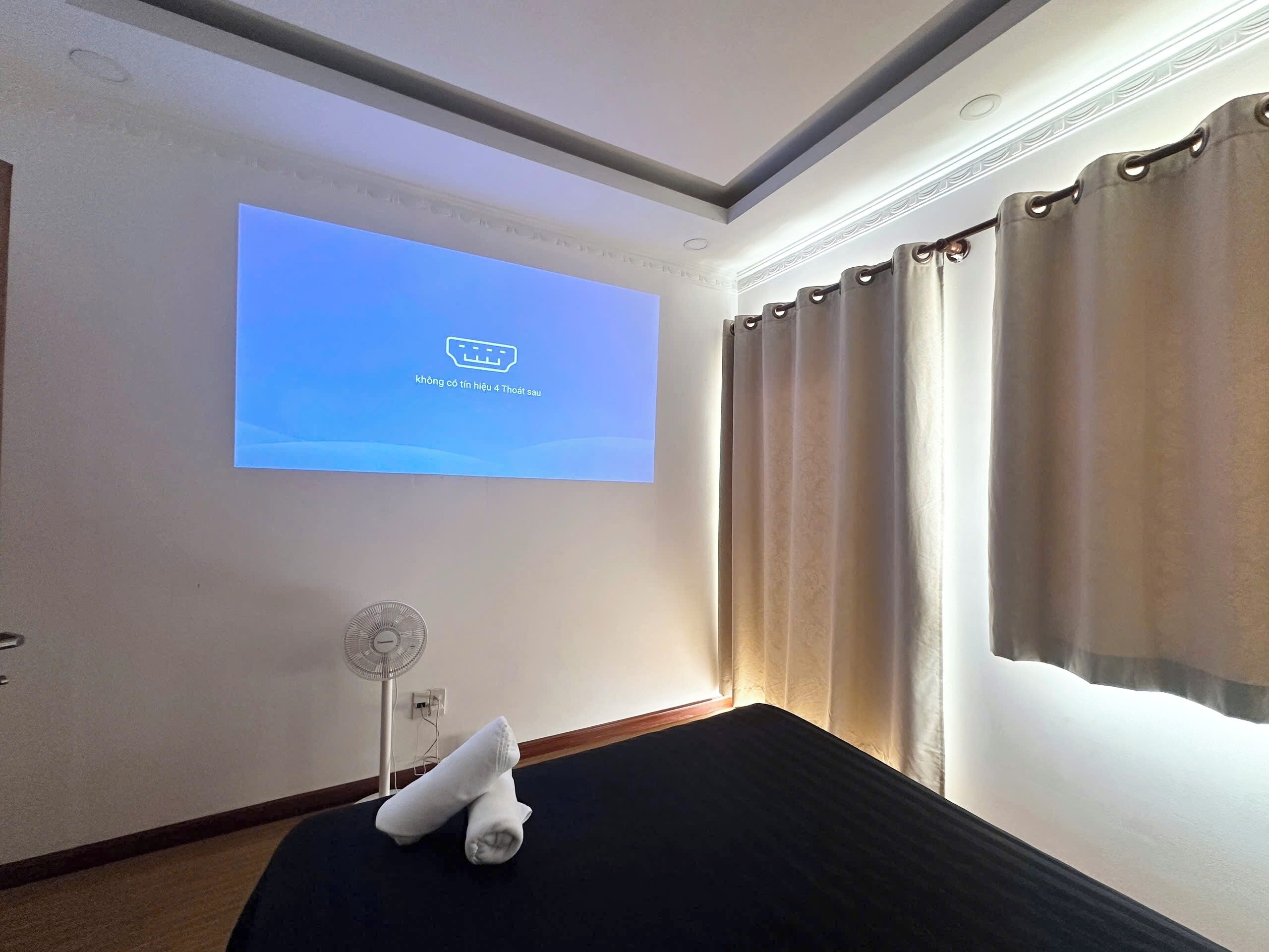 A hotel room with a bed, a wall-mounted projector, and a fan.