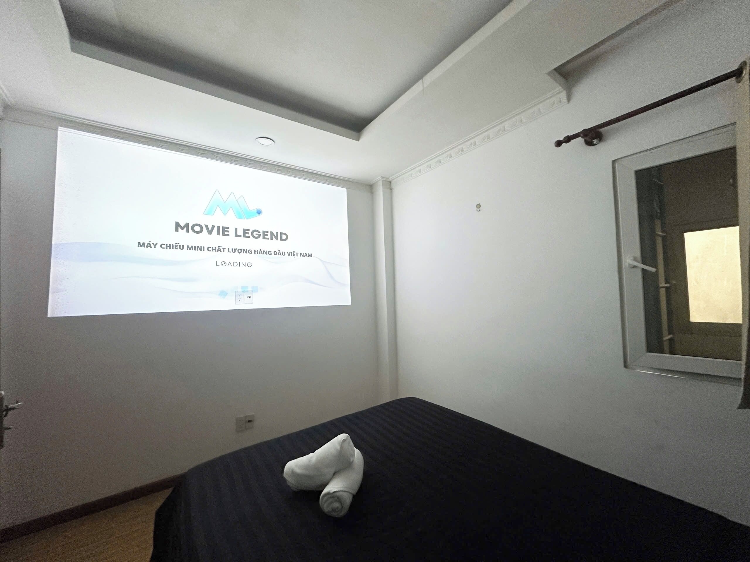 A hotel room with a bed, a projector screen displaying 'MOVIE LEGEND', and a window with a curtain.