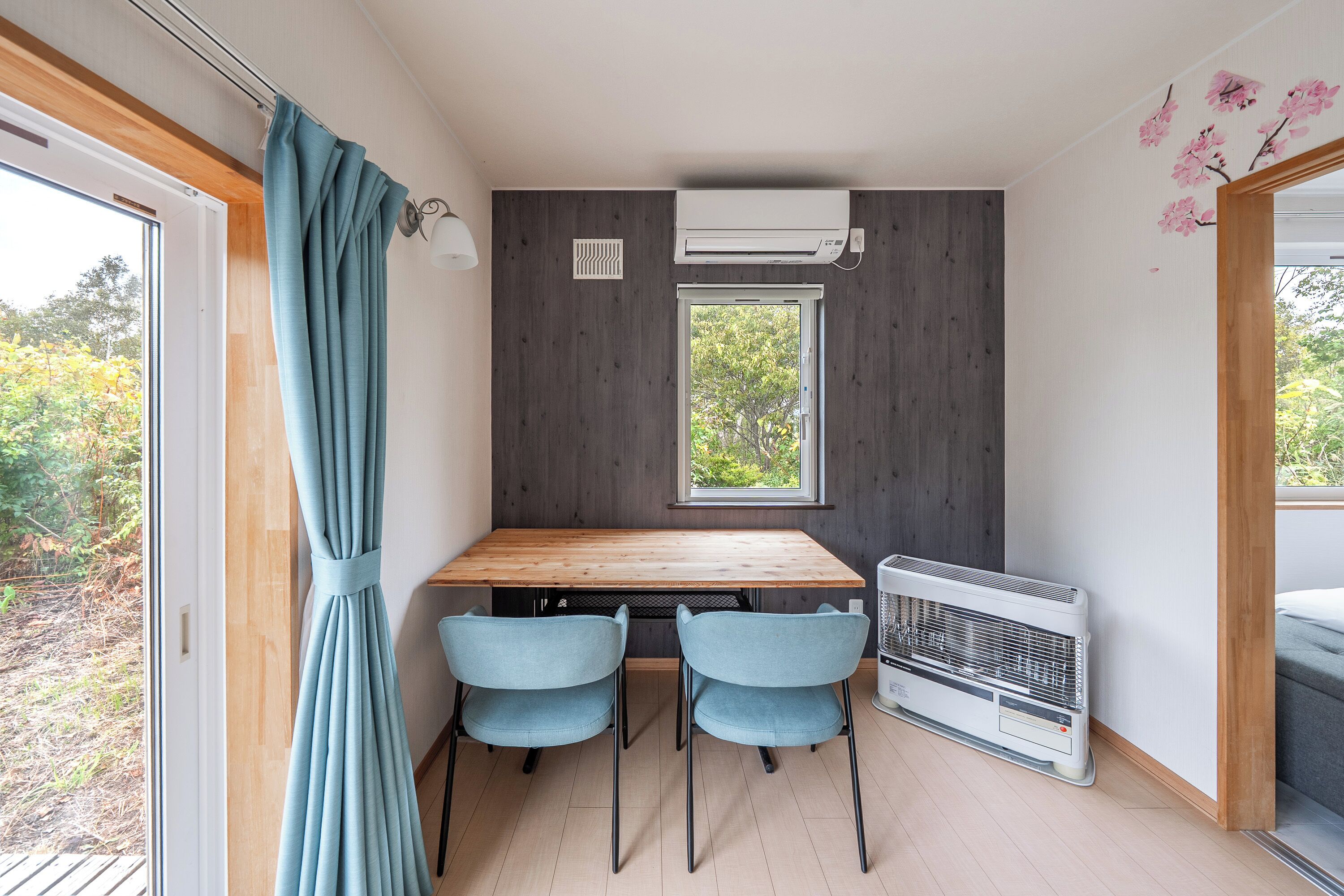 Une pièce de taille modeste, avec une table en bois, deux chaises, une fenêtre, un climatiseur et une vue sur la verdure à l’extérieur.