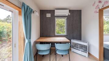 Une pièce de taille modeste, avec une table en bois, deux chaises, une fenêtre, un climatiseur et une vue sur la verdure à l’extérieur.