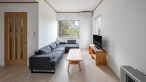 Un salon moderne avec un canapé d’angle, une table basse en bois, un téléviseur posé sur un meuble en bois et une grande fenêtre laissant entrer la lumière naturelle.