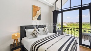 A modern bedroom with a large bed, a bedside table with a lamp, a painting on the wall, and a balcony with a view of greenery.