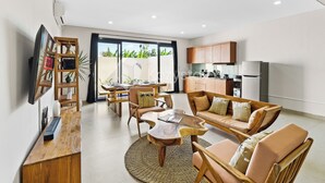 A modern living room with a wooden coffee table, sofa, and TV stand. There is a kitchen area with cabinets and appliances.