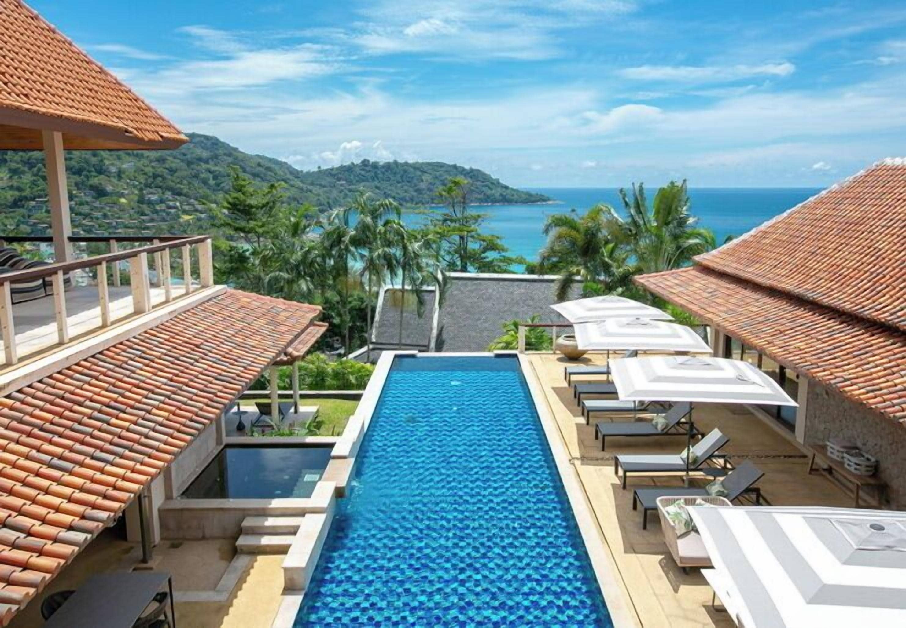 A swimming pool with lounge chairs and umbrellas, surrounded by buildings with red-tiled roofs, overlooking a mountainous landscape and a body of water.