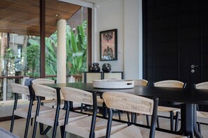 A dining area with a black table and wicker chairs, a large window with a view of greenery, and a black door. -  Katamanda Villa Allegra 5BR Seaview villa Kata (Karon)
