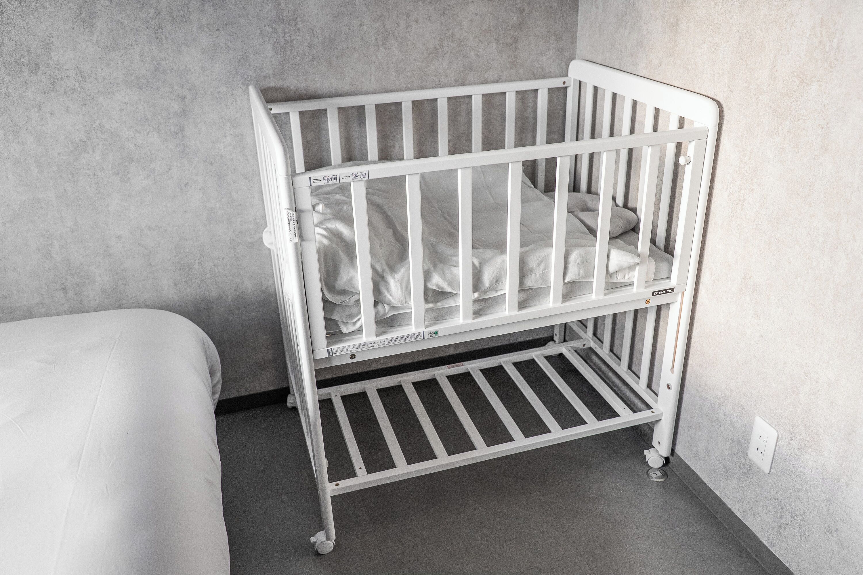 A white crib with a mattress and a lower shelf, positioned against a grey wall in a room with a grey floor.