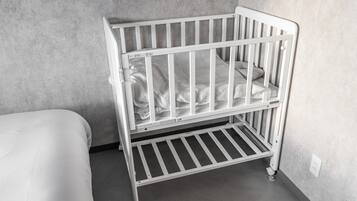 A white crib with a mattress and a lower shelf, positioned against a grey wall in a room with a grey floor.