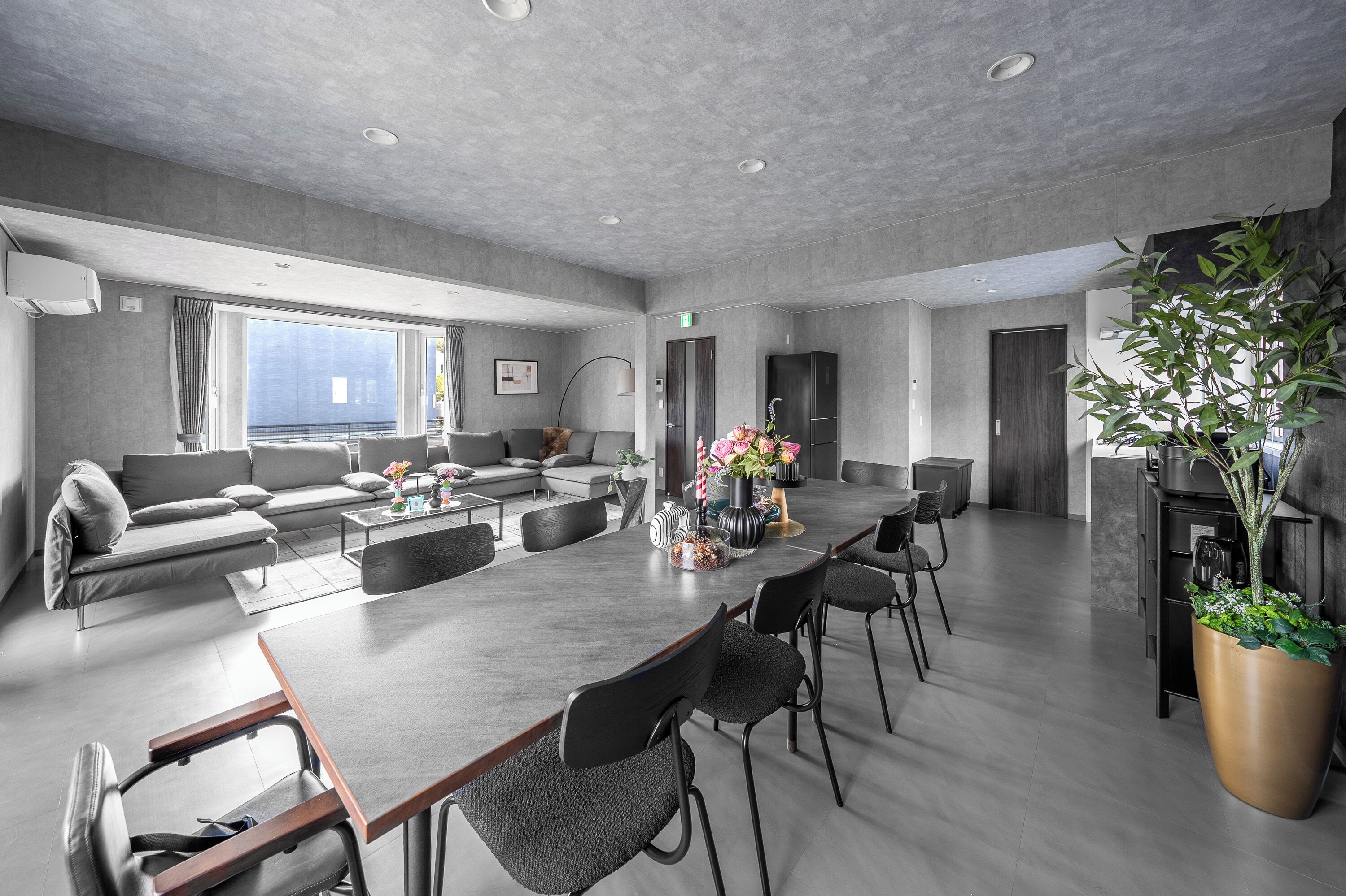 A spacious, modern hotel room with a large dining table, comfortable seating, a large window, and a potted plant.