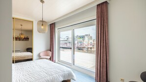 A modern bedroom with a large bed, a chair, a mirror, and a view of the city through a large window. - Beautiful penthouse flat with central location (Koksijde)