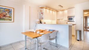 A modern kitchen with white cabinetry, a central island, and a dining area with white chairs. - Cosy flat with frontal sea view (Koksijde)