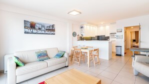 Un salon moderne avec un canapé, une table basse et un coin repas avec des chaises. Au fond, on aperçoit une cuisine avec des meubles blancs et un pare-feu en carrelage.