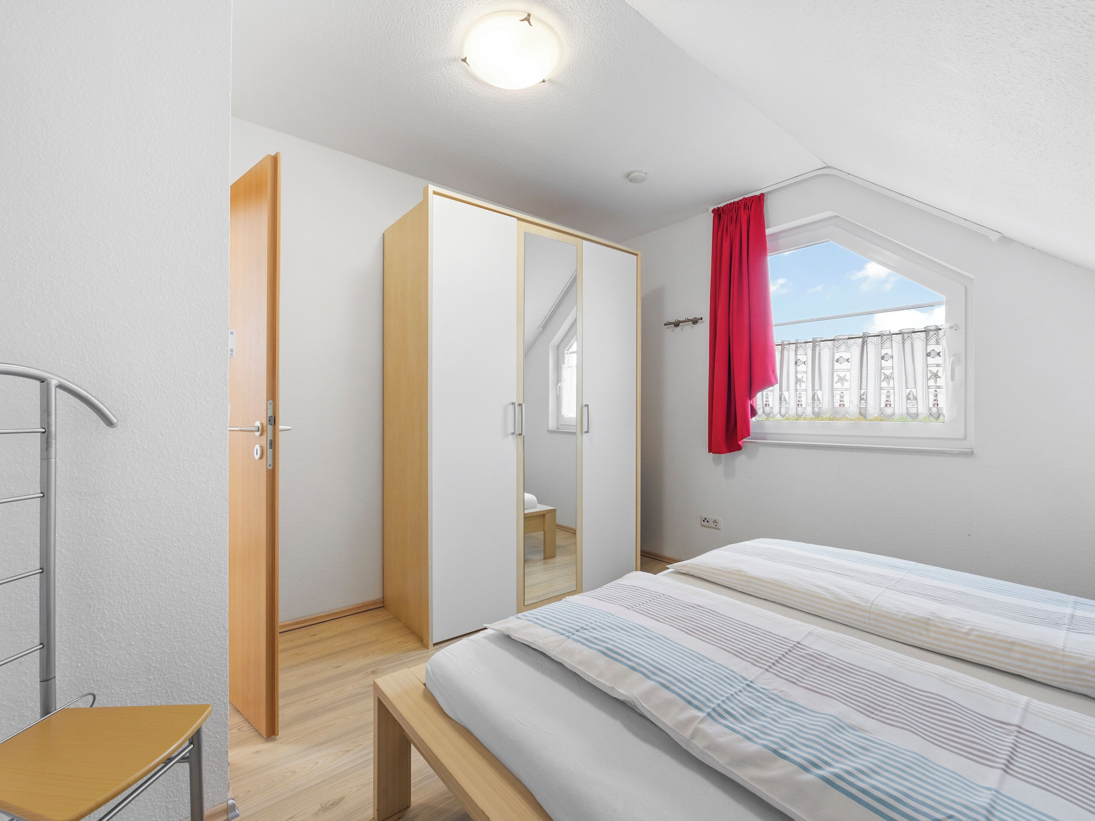 A bedroom with a bed, a wooden bench, a wardrobe, and a window with a red curtain.