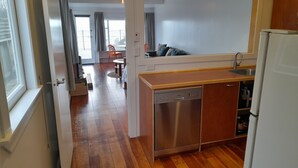 Une cuisine compacte équipée d’un lave-vaisselle, d’un évier et d’un réfrigérateur. On aperçoit un coin repas avec une table et des chaises à travers une porte ouverte.
