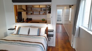 Une chambre à coucher comprenant un lit, des tables de chevet, un bureau intégré, un miroir et une vue sur un couloir menant à la cuisine.