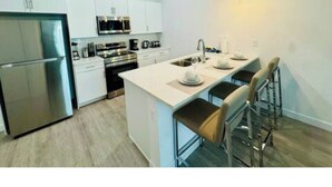 A modern kitchen with white cabinetry, stainless steel appliances, and a central island with bar stools. - The Sun-Kissed Retreat / near Disney (Davenport)