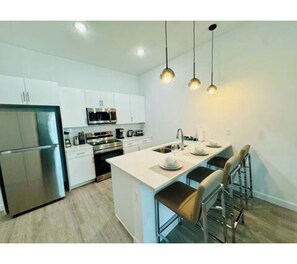 A modern kitchen with white cabinetry, stainless steel appliances, and a central island with bar stools. - The Sun-Kissed Retreat / near Disney (Davenport)
