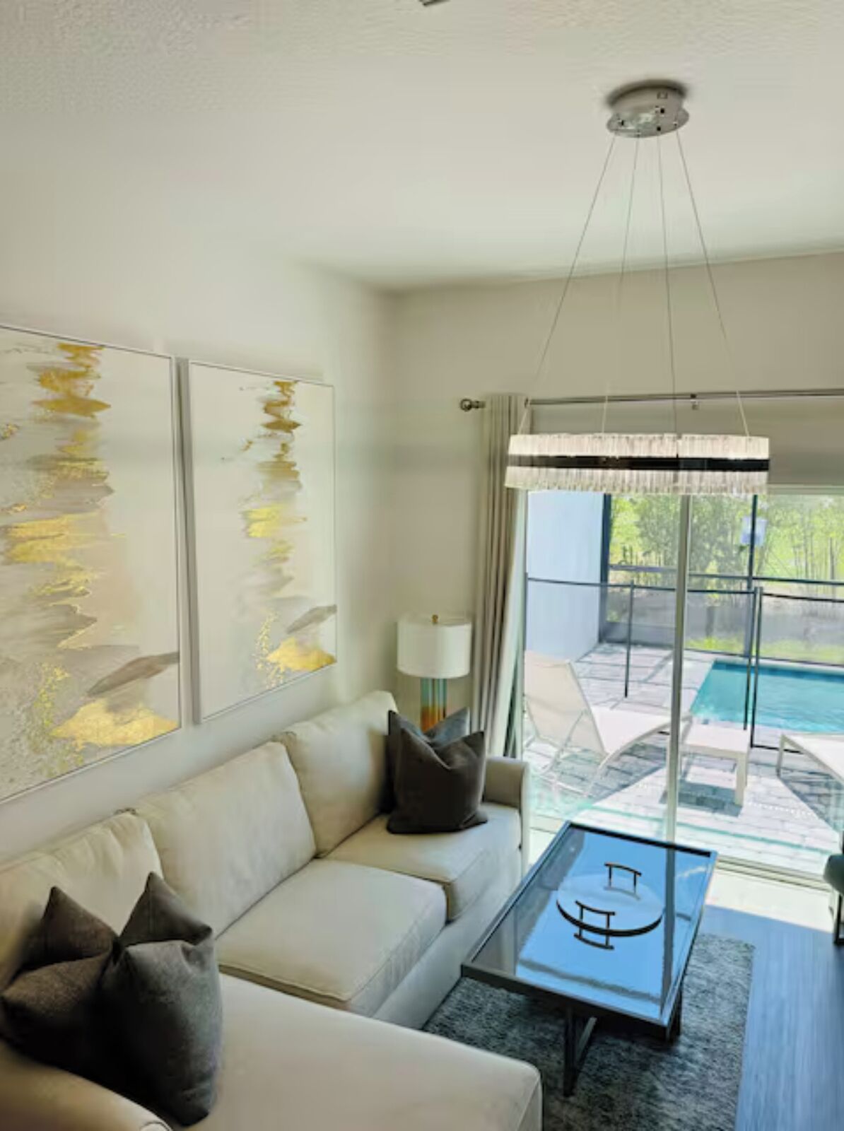 Una sala de estar moderna con un sofá, una mesa de centro y arte abstracto en la pared. Se ve una ventana grande con vistas a una piscina y vegetación.