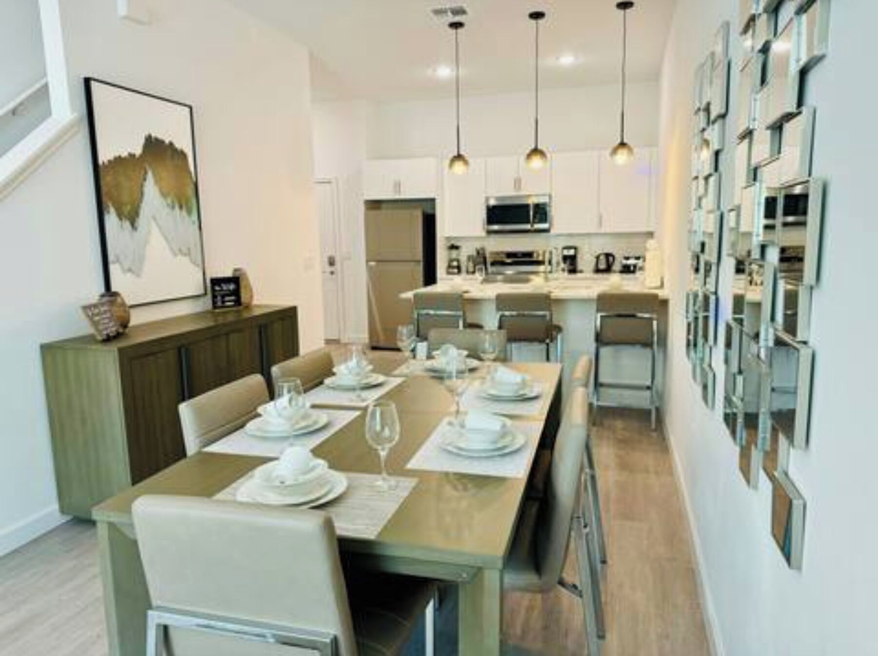 A modern dining area with a long table set for four, a kitchen in the background, and pendant lights above.