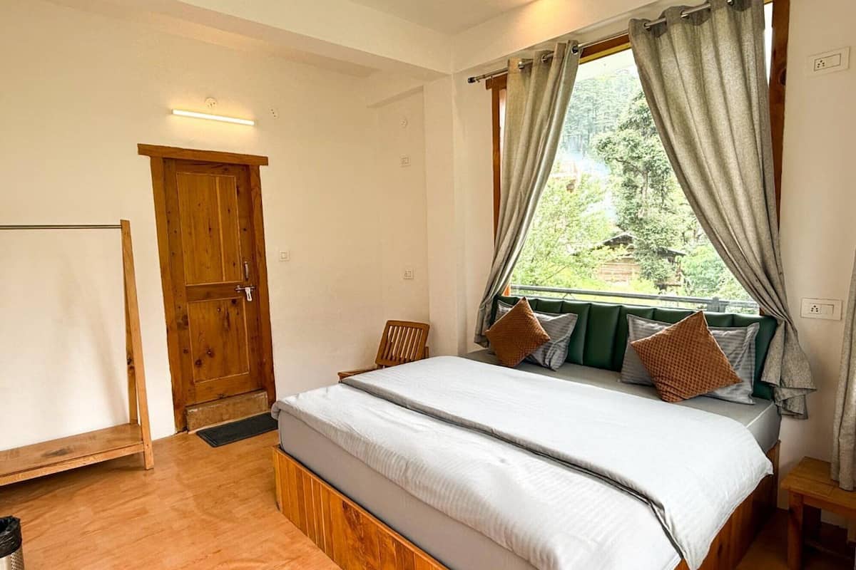 Quarto com cama de madeira, janela com cortinas, um banco estofado verde e porta de madeira.