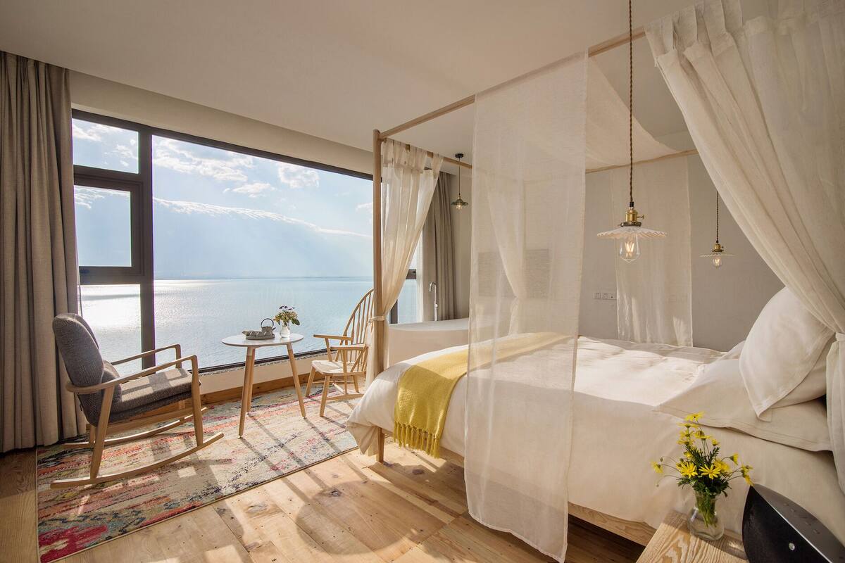 A bedroom with a large window offering a view of the sea, a bed with a canopy, a small table with a vase, and a chair.