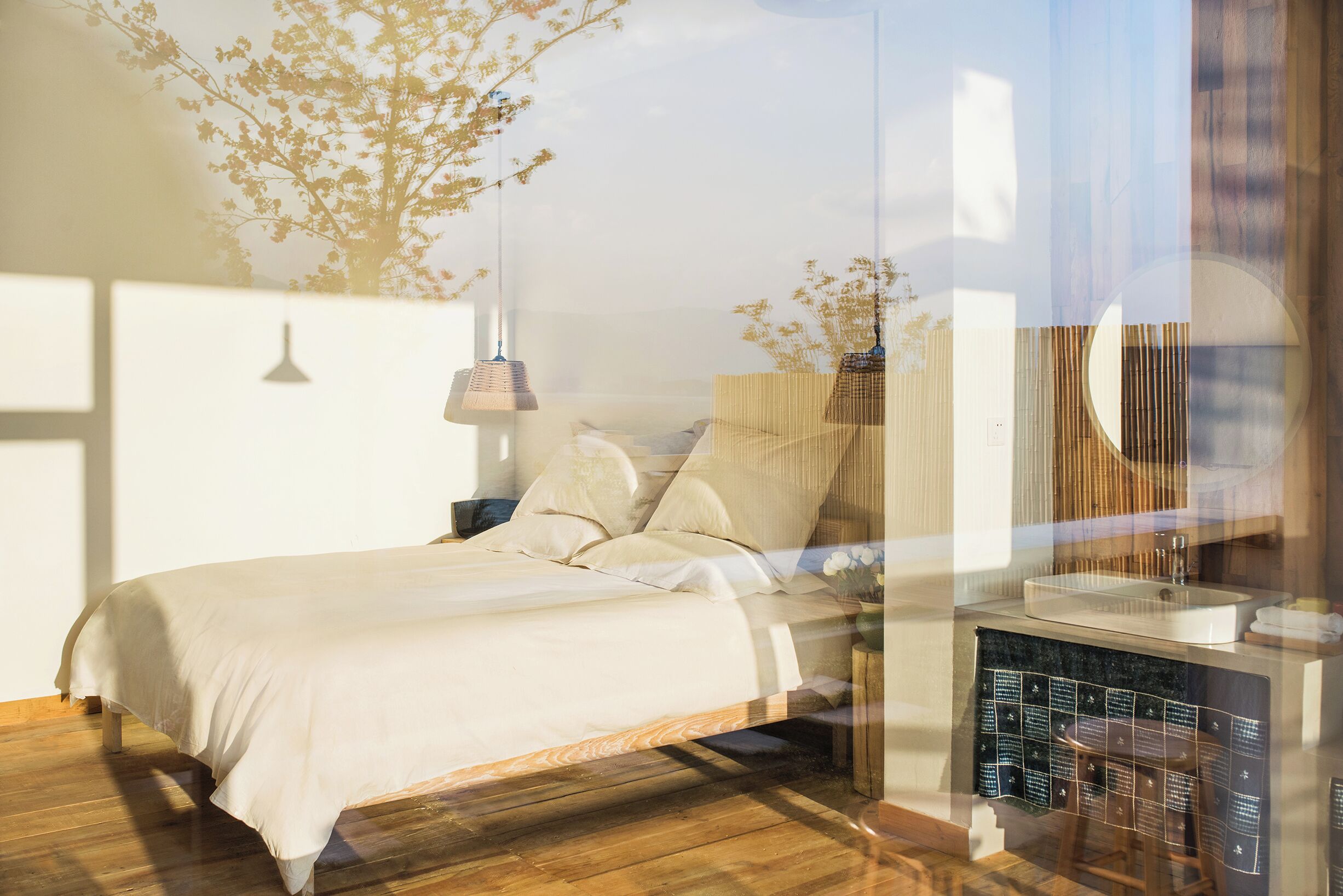 A bedroom with a wooden floor, a bed with white bedding, a nightstand, a wall-mounted lamp, and a window with a view of trees.