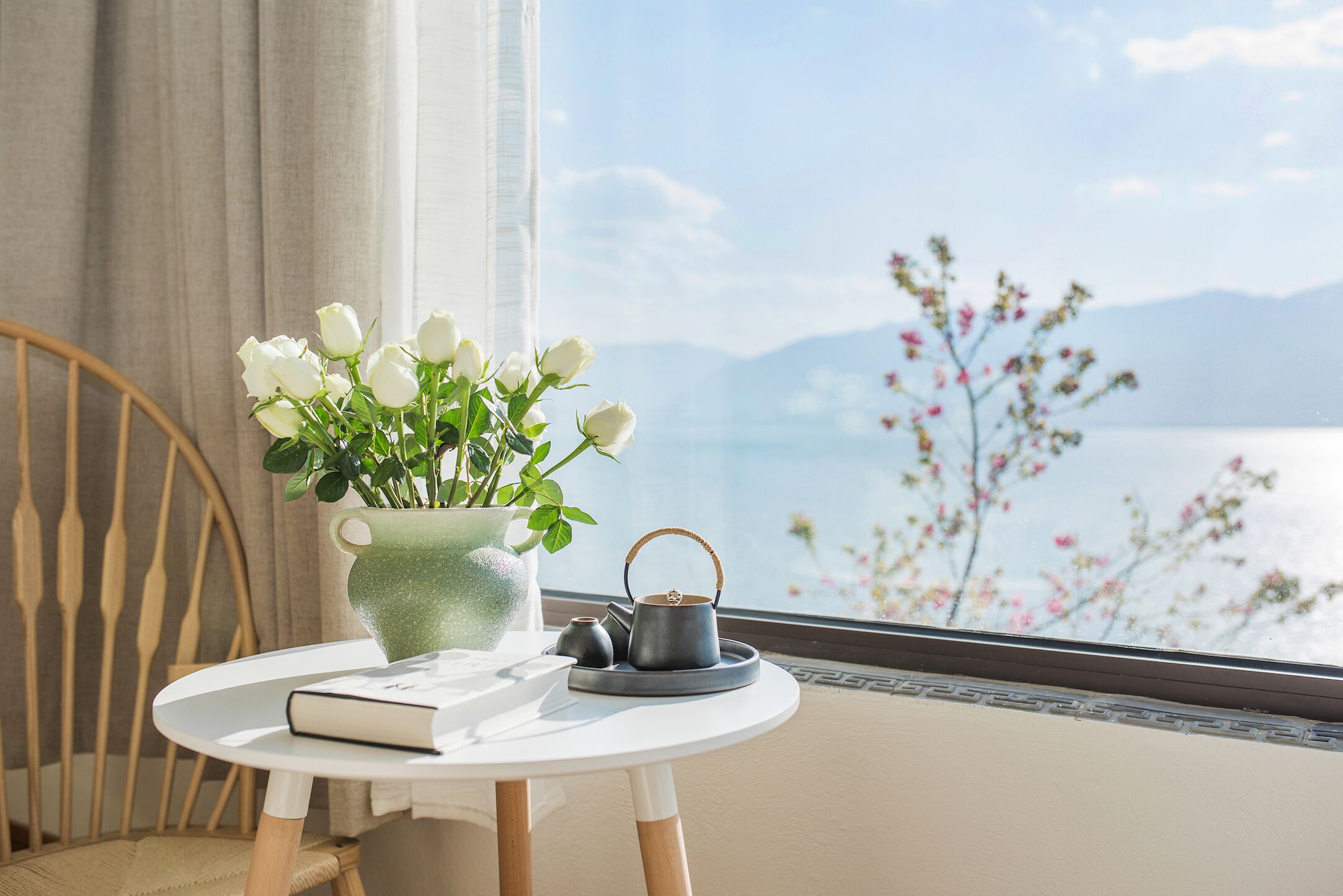 A round table with a book, a green vase with white flowers, and a black teapot on a tray.