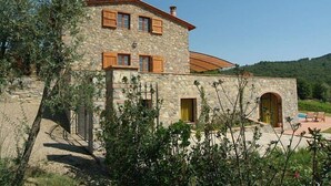Un edificio de piedra con contraventanas de madera, una zona de piscina y un paisaje montañoso de fondo.