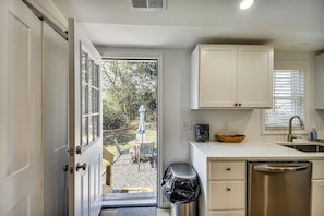 Una cocina con gabinetes blancos, lavavajillas de acero inoxidable y vistas a un espacio de estar exterior a través de la puerta abierta.