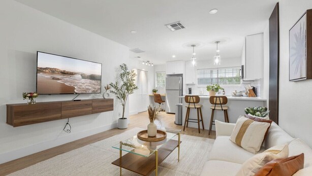 A modern living room with a flat-screen TV, a coffee table, and a sofa. There is a kitchen area with a refrigerator, a sink, and a window.