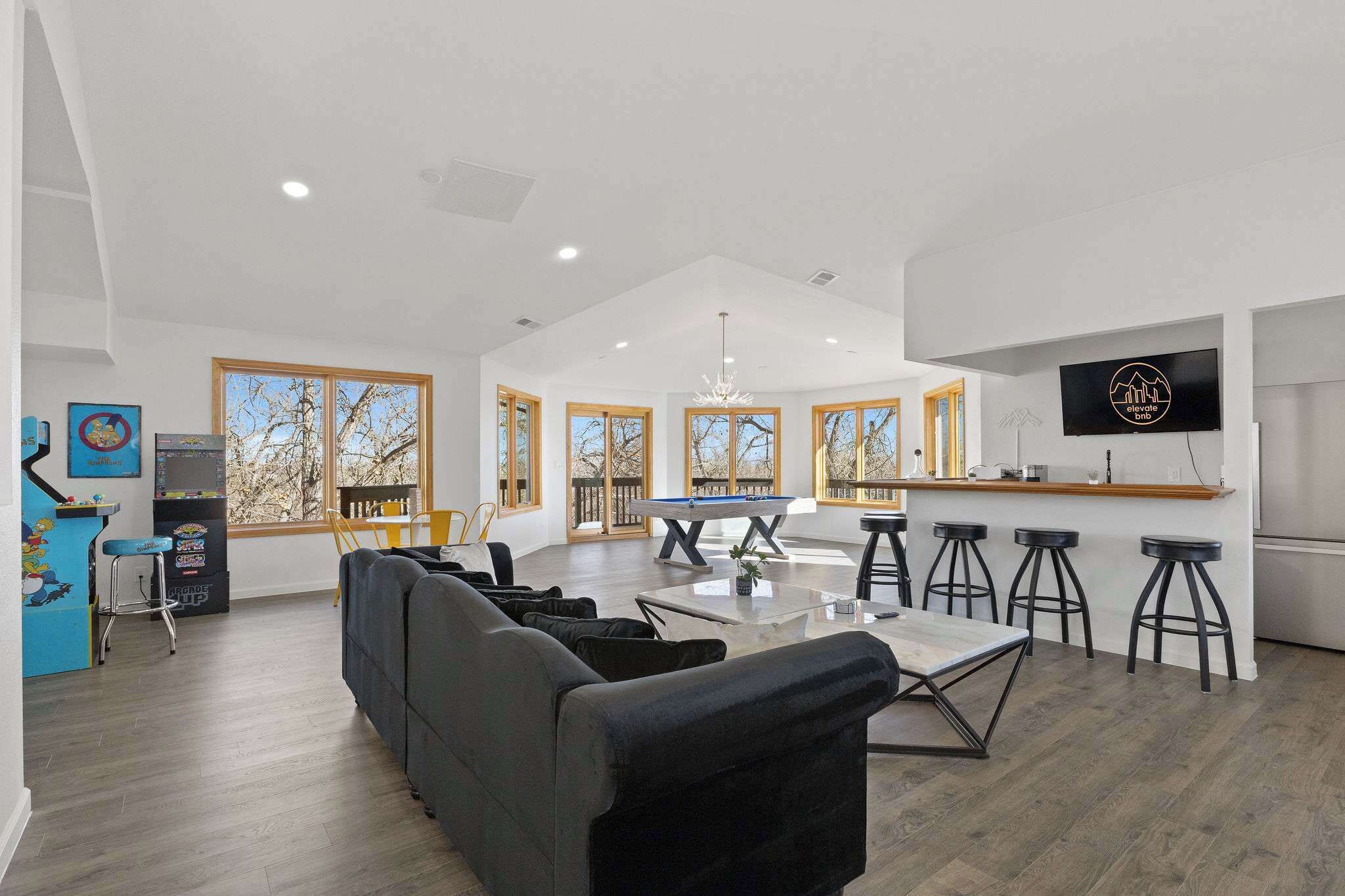 A spacious living area with a sofa, dining table, and bar stools. There is a pool table, a TV mounted on the wall, and large windows with a view of trees.