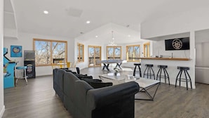 A spacious living area with a sofa, dining table, and bar stools. There is a pool table, a TV mounted on the wall, and large windows with a view of trees. - Ultimate Treehouse Mansion w/ Swim Spa, Sleeps 31 (Lakewood)