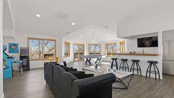 A spacious living area with a sofa, dining table, and bar stools. There is a pool table, a TV mounted on the wall, and large windows with a view of trees.