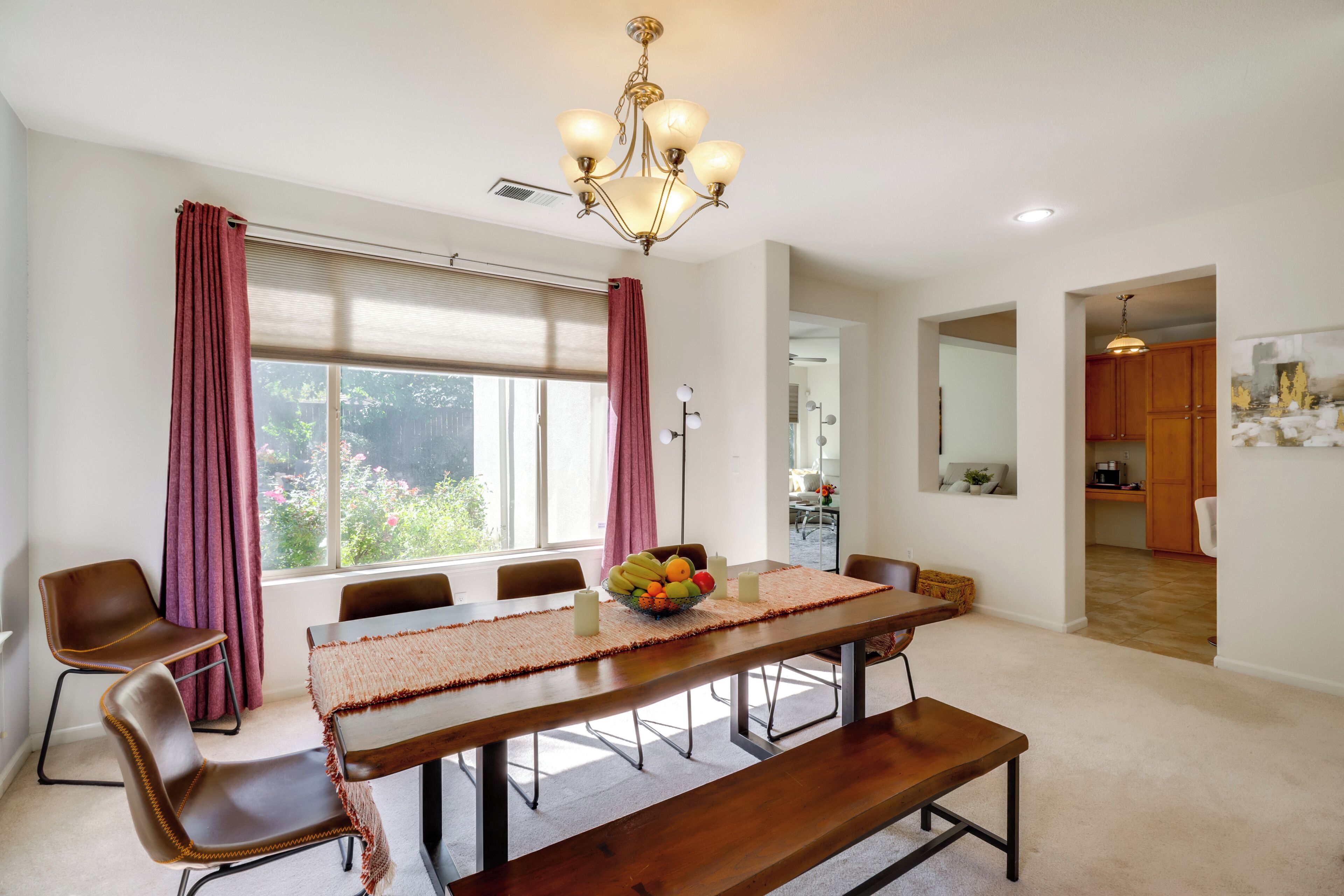 Un espace repas comprenant une longue table en bois, six chaises, un lustre et une fenêtre avec des rideaux.