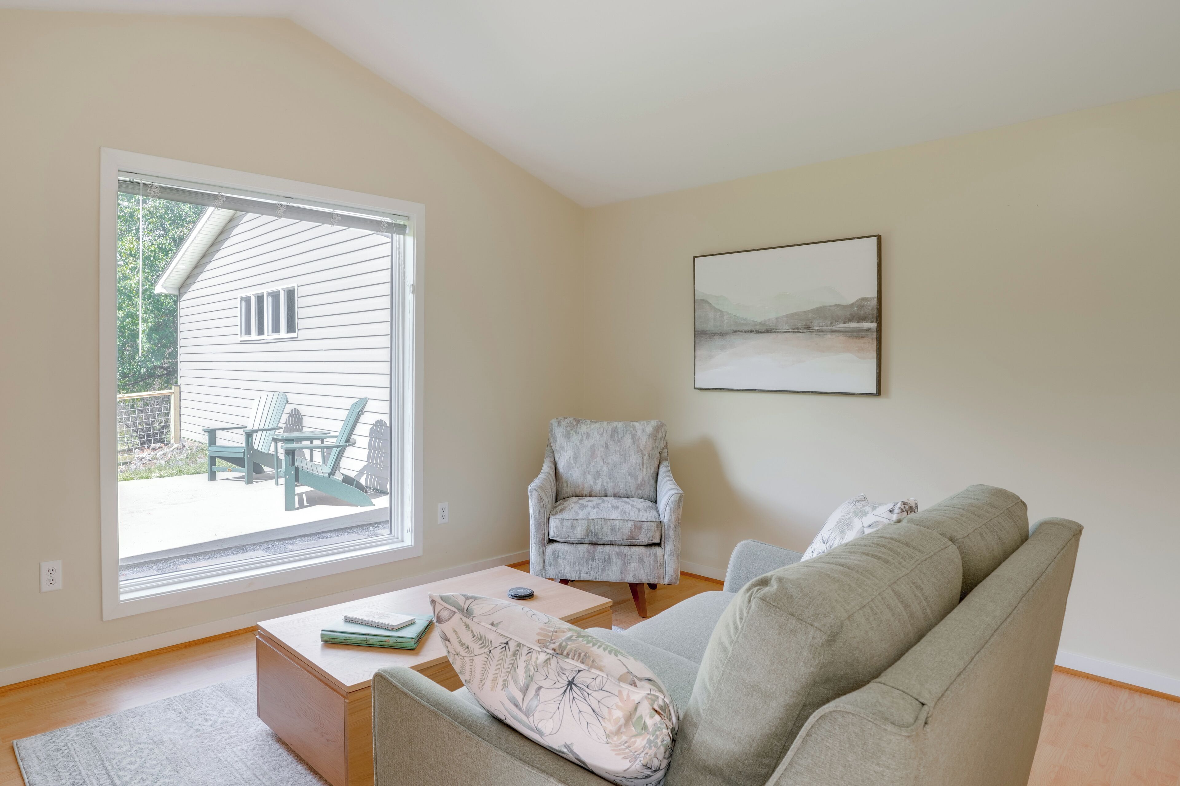 Un salon avec une grande fenêtre, un fauteuil gris, un canapé gris et une petite table sur laquelle sont posés des livres et un coussin décoratif.