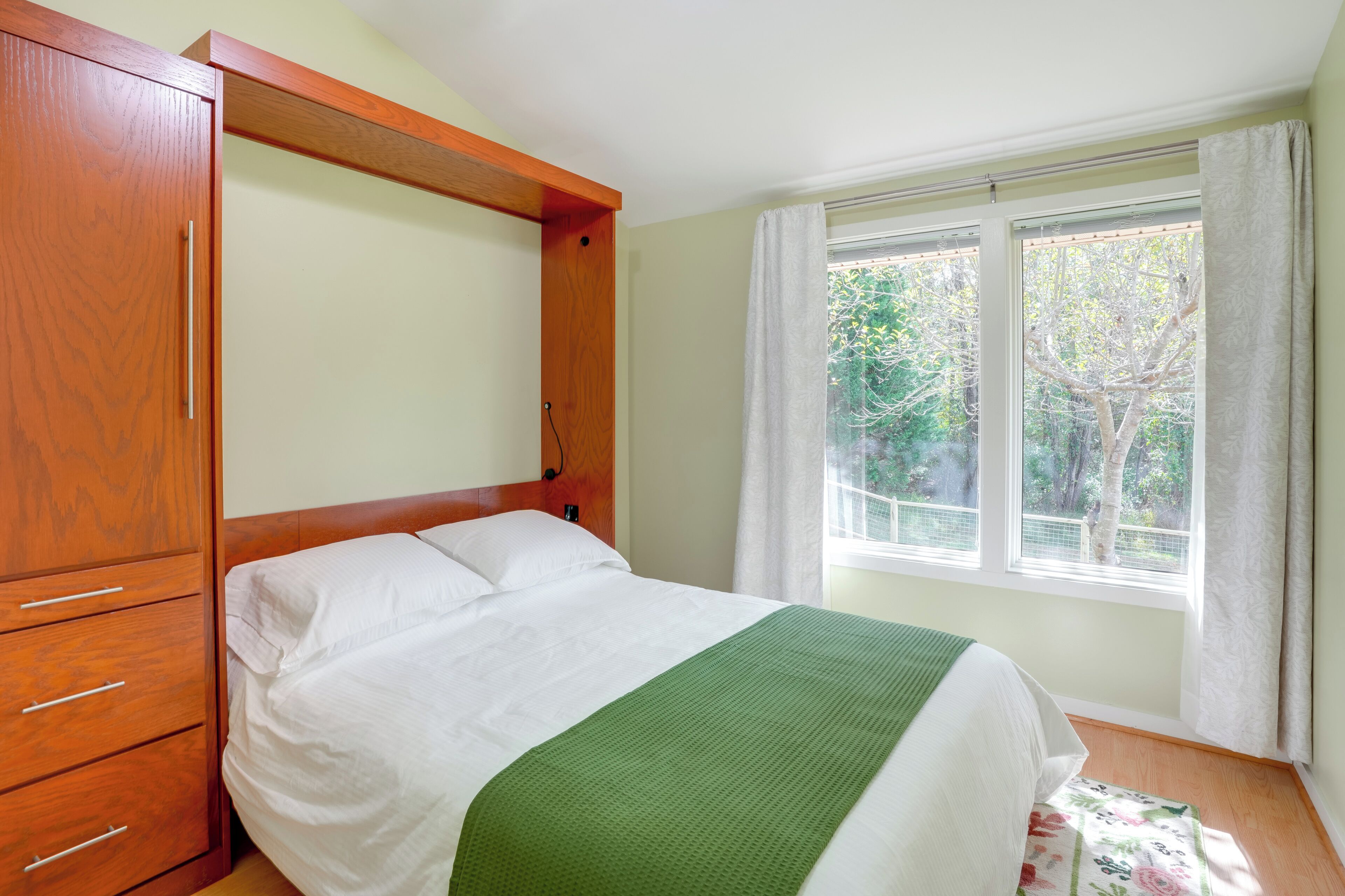 Een slaapkamer met een houten garderobe, een bed met wit beddengoed en een groene plaid, en een raam met uitzicht op bomen.