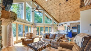A spacious living room with a wooden ceiling, large windows, a sofa, a coffee table, and a kitchen area visible through the glass doors. - Secluded Lake Lanier Cabin w/ Game Area (Gainesville)