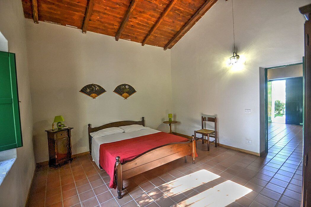 Quarto com cama de madeira, duas mesas de cabeceira, uma cadeira e um ventilador na parede.