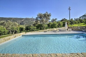 Une piscine rectangulaire à l’eau d’un bleu limpide, entourée d’un mur de pierre et bordée d’une pelouse parsemée d’arbres, avec une colline en arrière-plan.