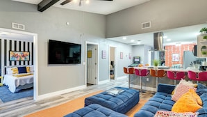 Un salon moderne avec un canapé d’angle bleu, un tapis orange et un coin cuisine doté d’un mur en briques rouges et de chaises orange.