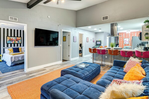 A modern living room with a blue sectional sofa, an orange rug, and a kitchen area with a red brick wall and orange chairs. - 9 Mi to Legoland: Family Gem w/ Games Galore! (Winter Haven)