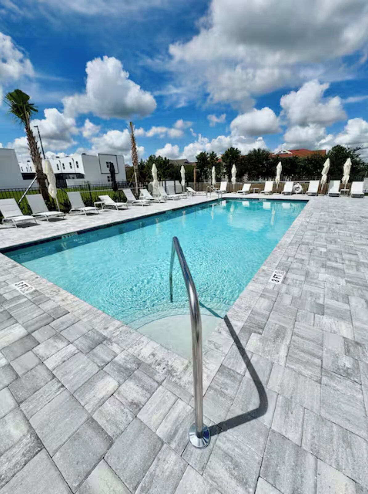 Piscina rectangular con barandilla de seguridad metálica, rodeada de sillas de descanso y sombrillas blancas, bajo un cielo azul despejado con nubes.