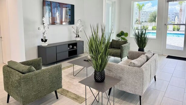 A modern living room with a grey sofa, a green armchair, a coffee table, and a TV on the wall. There are potted plants and a view of a swimming pool outside. - The Villa Retreat / near Disney (Davenport)