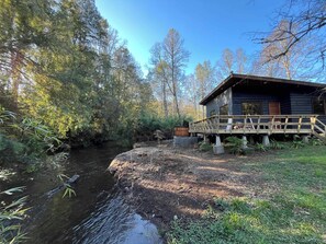 A cabin with a wooden deck overlooking a river, surrounded by trees.