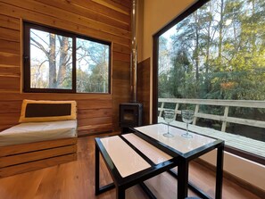A wooden cabin interior with a sofa, a table, and a large window offering a view of trees.