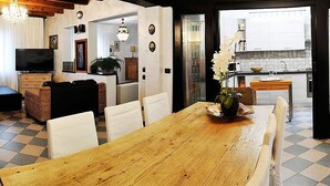 A dining area with a wooden table and white chairs, a television in the background, and a kitchen visible through an open door. - Villa Bacio: An elegant and welcoming two-story villa situated at a short distance from the Adige river, with Free WI-FI. (Castelbaldo)