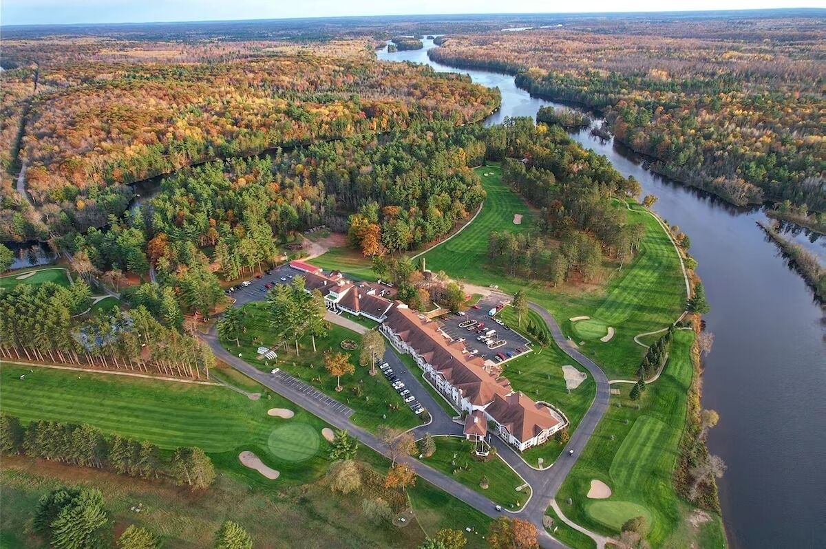 An aerial view of a golf course with a clubhouse, surrounded by dense forests and a river.